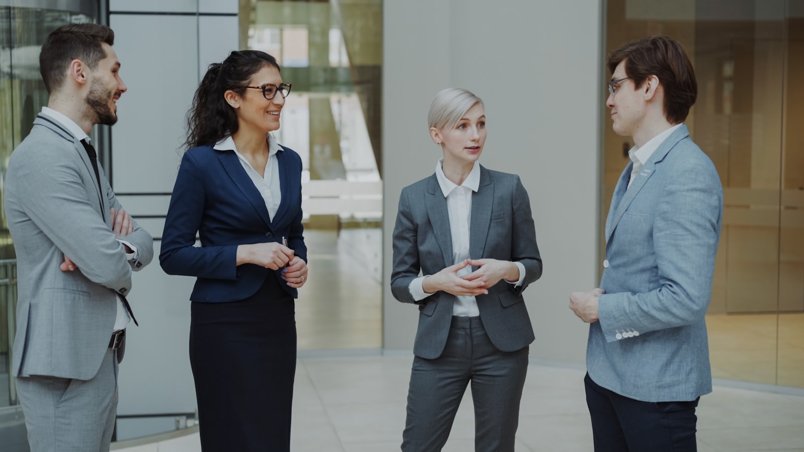 Quatre professionnels en tenue de bureau échangeant debout dans le hall vitré d'un immeuble d'entreprise.
